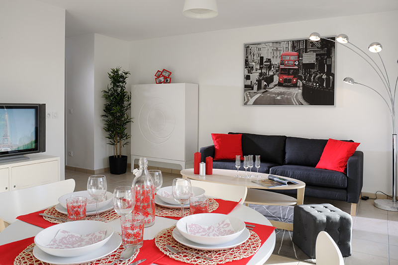 Pièce de vie avec canapé gris au fond, meuble haut blanc et devant une table ronde dressée pour 4 en rouge et blanc